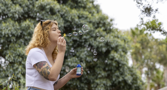 A woman blows bubbles with a bubble wand, there are green trees in the background