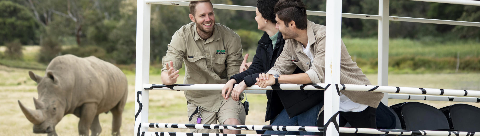 A zoo keeper stands on the back of a vehicle talking to two people. A rhino can be seen in the background.