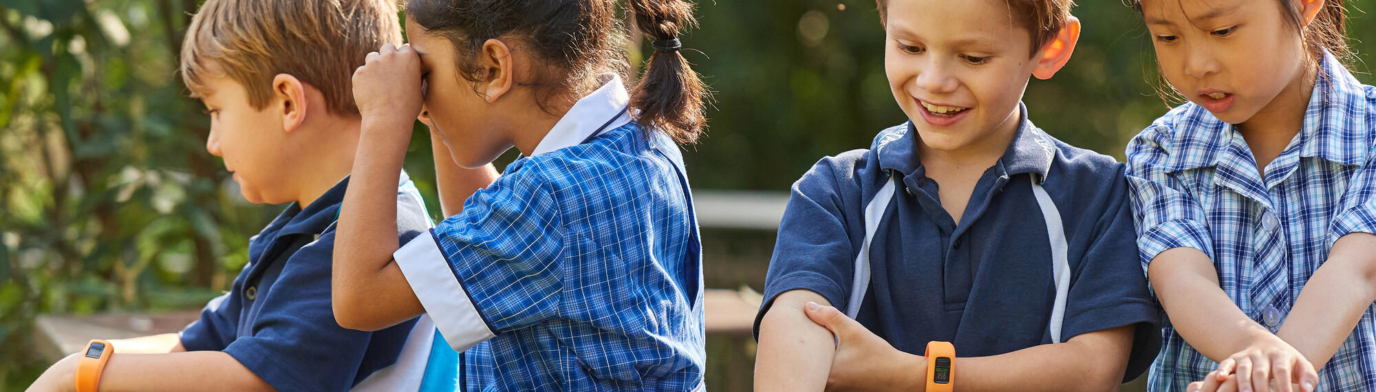 A group of young, primary school aged children sit and play, all dressed in blue uniforms and two wearing orange-banded watches.