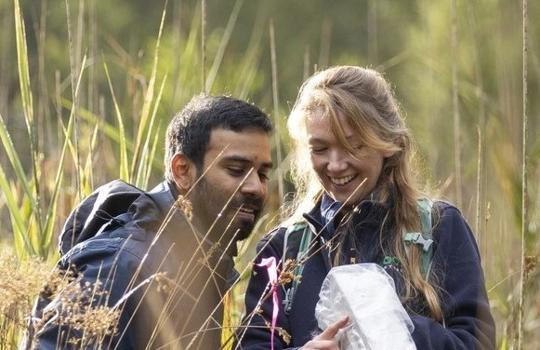 Two field scientists sit in grasslands and stare into a small bag with smiles on their faces