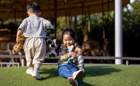 Two young guests holding plush toys, as one looks to the camera smiling and another faces away toward the Carousel.
