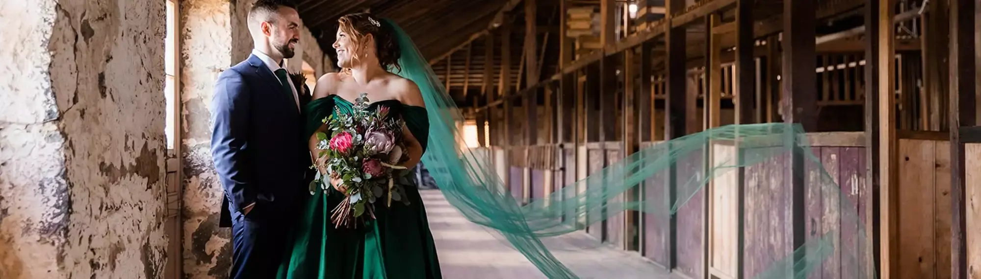 A man and woman dressed in formal wedding wear are standing in a rustic room, happily looking at each other.