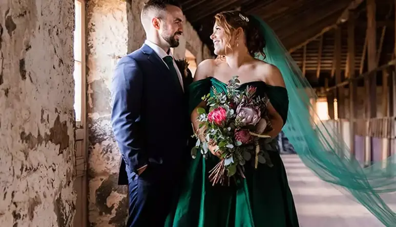A man and woman dressed in formal wedding wear are standing in a rustic room, happily looking at each other.