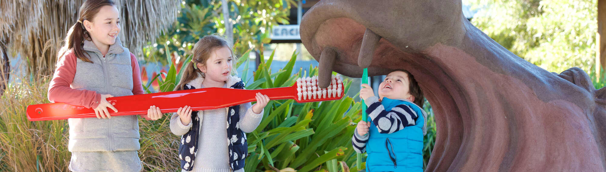 Three children brushing fake Hippo's tooth with a giant red toothbrush.