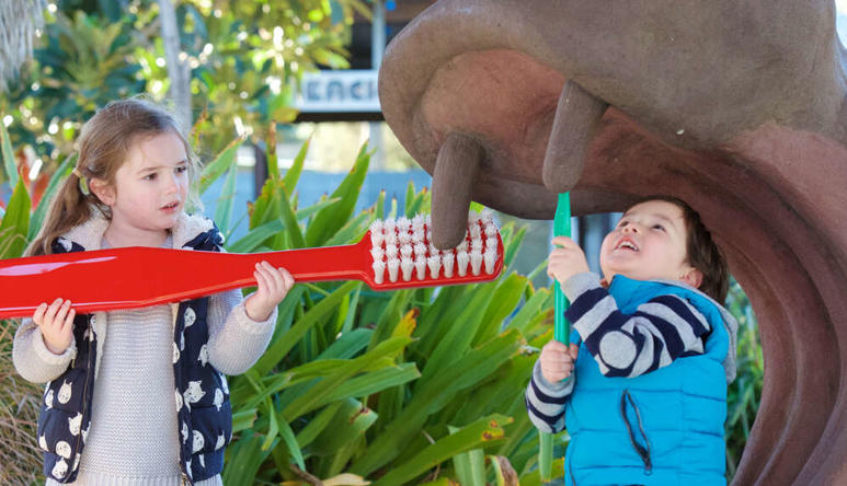 Three children brushing fake Hippo's tooth with a giant red toothbrush.