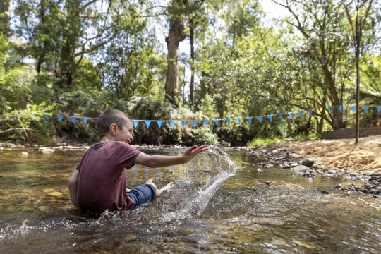 A young guest joyfully slinging water in the shallow Poorneet creek, turned away from the camera and looking right.