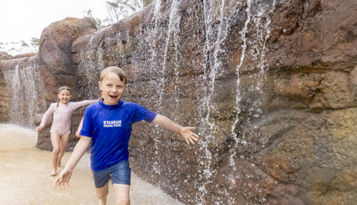 Three young guests run past a small waterfall, running towards and smiling to the camera, with their left arms stretched out to catch water.