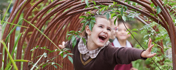 Girl in brown school uniform smiles as she walks through an archway and two other students can be seen behind her.