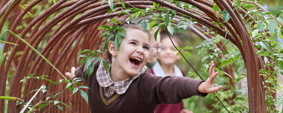Girl in brown school uniform smiles as she walks through an archway and two other students can be seen behind her.