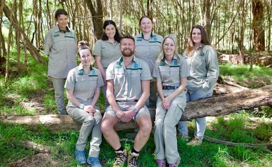 A group of seven Zoo employees sit on and stand behind a log, in a group while smiling.