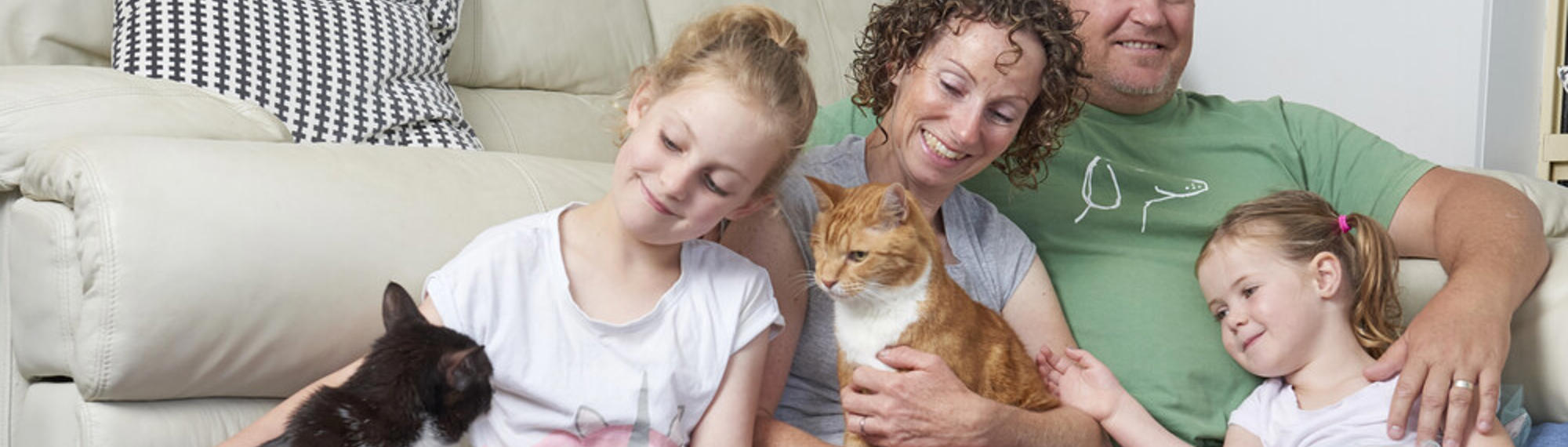 A family of four sitting in front of a couch patting two cats and smiling.