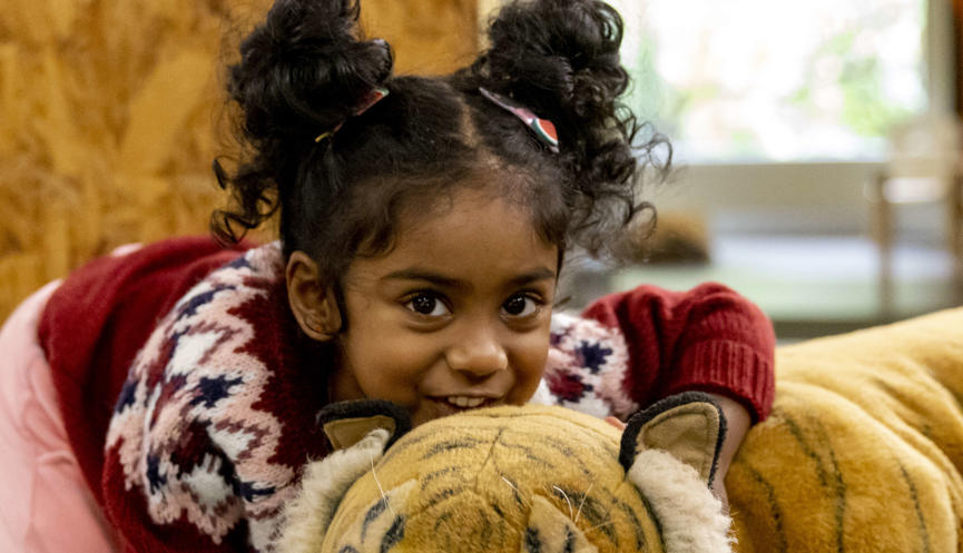 A young guest cuddles the large plush Tiger of Keeper Kids, while she faces the camera, crouching and smiling.