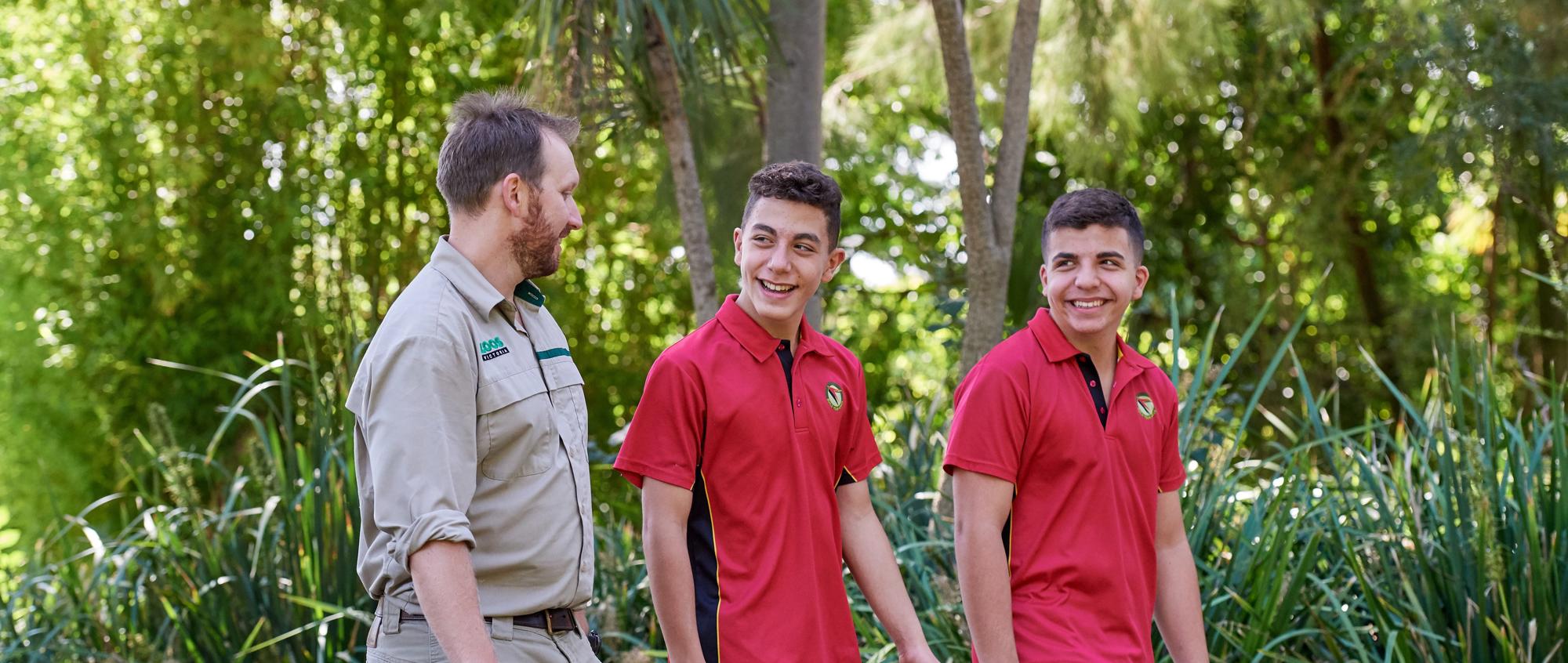 Two middle-high school aged teens in red school uniforms, walk alongside a Zoo employee dressed in khaki.