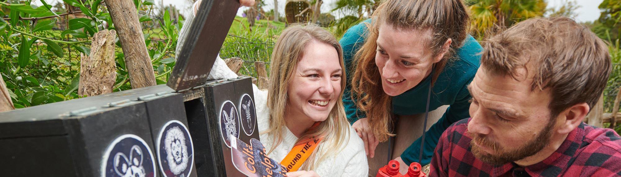 Two women laughing and a man participating in Race Around The Zoo retrieve clue cards from wooden boxes with animal faces painted on the front. 