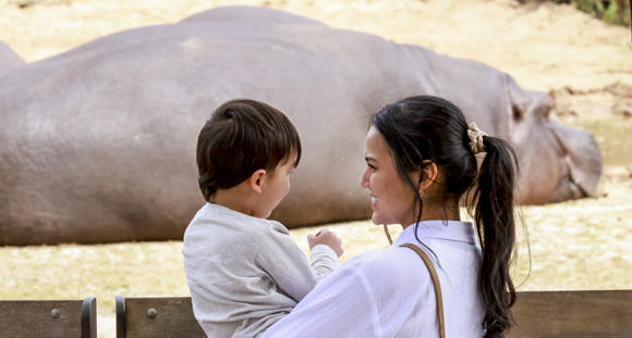 Two guests, mother on right holding up son on left, face each-other as they stand before a prone Hippo in the background, who is facing right and away from the camera.