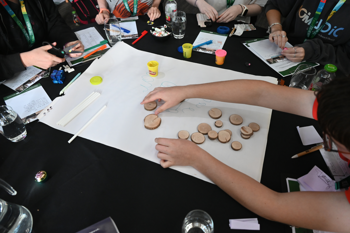 Five guests gathered around a black table, all engaged in a craft activity with paper, scissors, markers, three tubs of modelling clay and fifteen small wooden discs