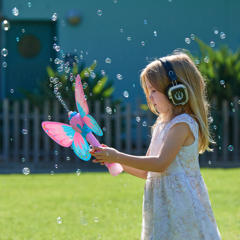 A young guest, wearing ear-muffs and holding a bubble blower with butterfly wings, as she stares at it and bubbles float around her.