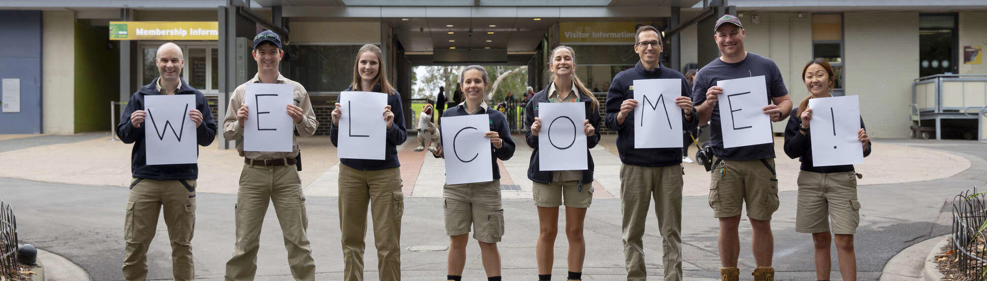 Eight Zoo staff each holding signs that spell out "W-E-L-C-O-M-E-(exclamation point!)."
