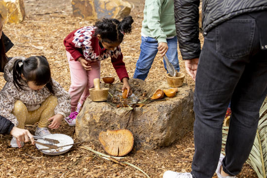 Two young guests make "dishes" from the "Mud Kitchen," accompanied by two adults supervising them.