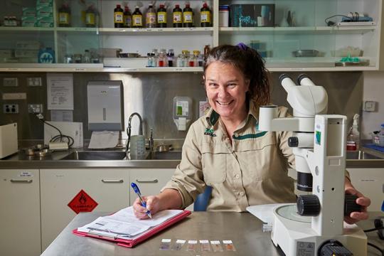 A smiling Vet, dressed in khaki, sits in front of a microscope with a clipboard and pen in her hand.