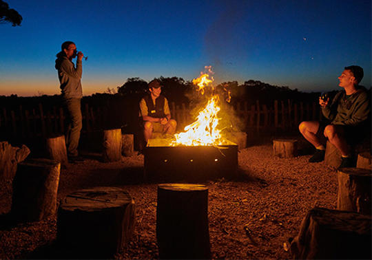 Two guests and a Keeper around a burning fire-pit at late dusk.