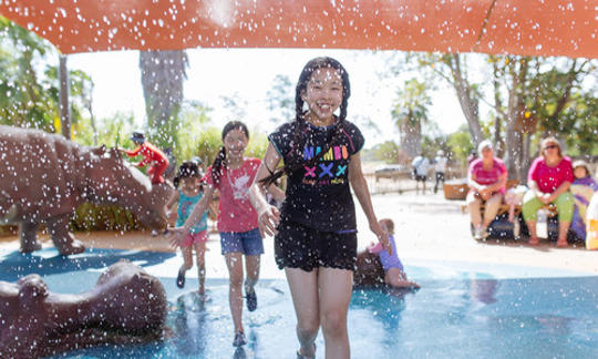 A young girl running through a fountain with water spraying her. Hippo statues in background.