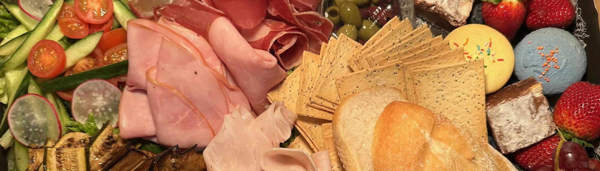 Hamper of assorted foods, including ham, turkey, cheese, zucchini, pastrami, tomato, olives, bread, cherries, strawberries, cookies and macarons.