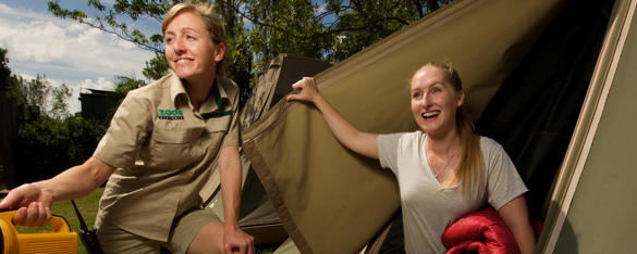 A woman lifts the opening of a tent and smiles, as a Zoo staff member squats next to her holding a torch.