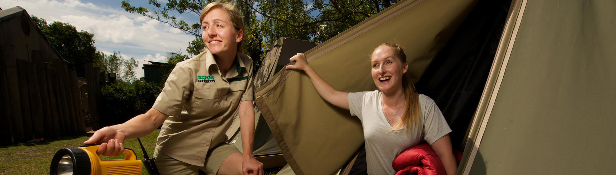 A woman lifts the opening of a tent and smiles, as a Zoo staff member squats next to her holding a torch.