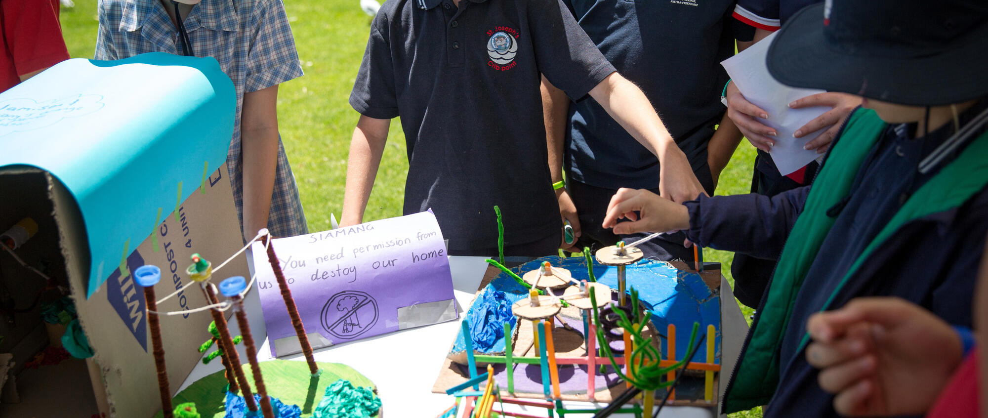 A group of primary school students wearing uniform, outside with a craft activity on a table; There is a handwritten sign on the table reading 'SIAMANG, You need permission from us to destroy our home' on it with an illustration of a tree with a cross through it.