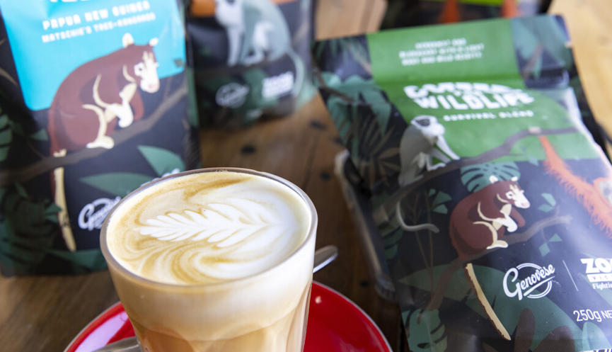 A milky coffee in a glass with latte art in the froth; the coffee sits on a saucer with a teaspoon, in front of bags of coffee on a table.