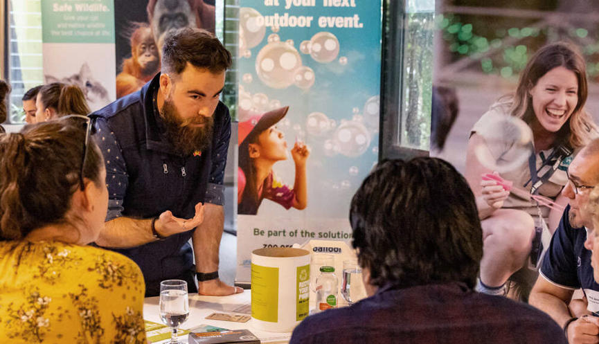 A group of four visitors sit around a table learning from a Zoos Victoria staff member, with "Blow Bubbles" posters beside them.