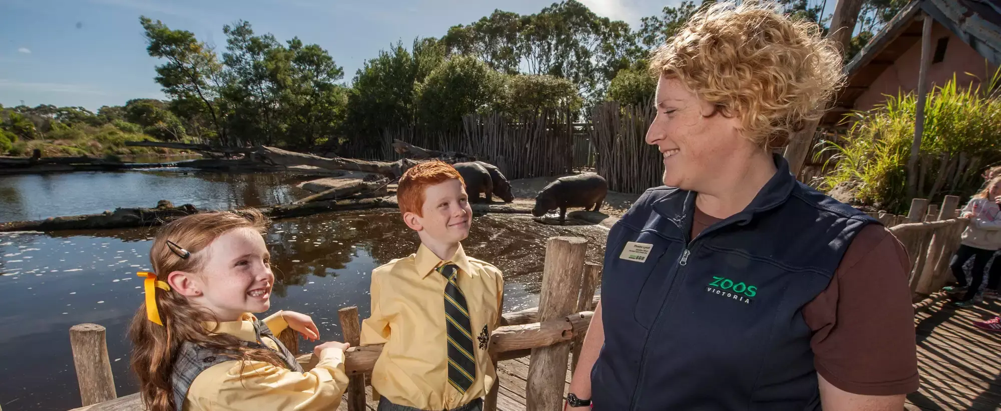 An Education Keeper, smiling to two students who are smiling back to her, on the Hippo Boardwalk with two Hippo's behind them.