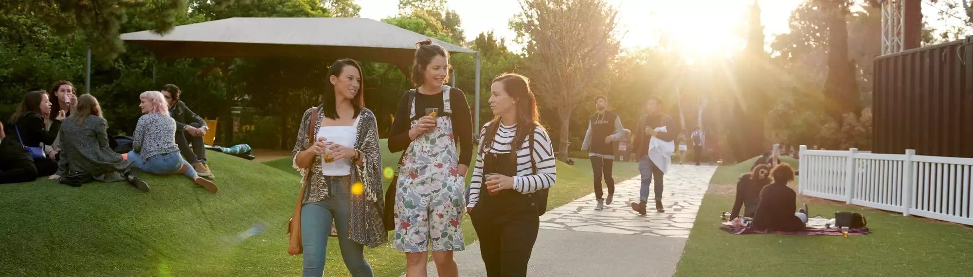 Three guests, holding drinks and talking to each-other, in the Carousel Park at dusk, seen with fifteen other guests.