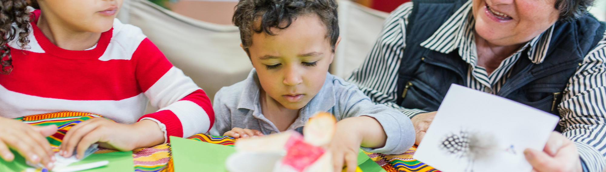 Two young guests making craft with paper and markers, joined by an elder woman on the right.