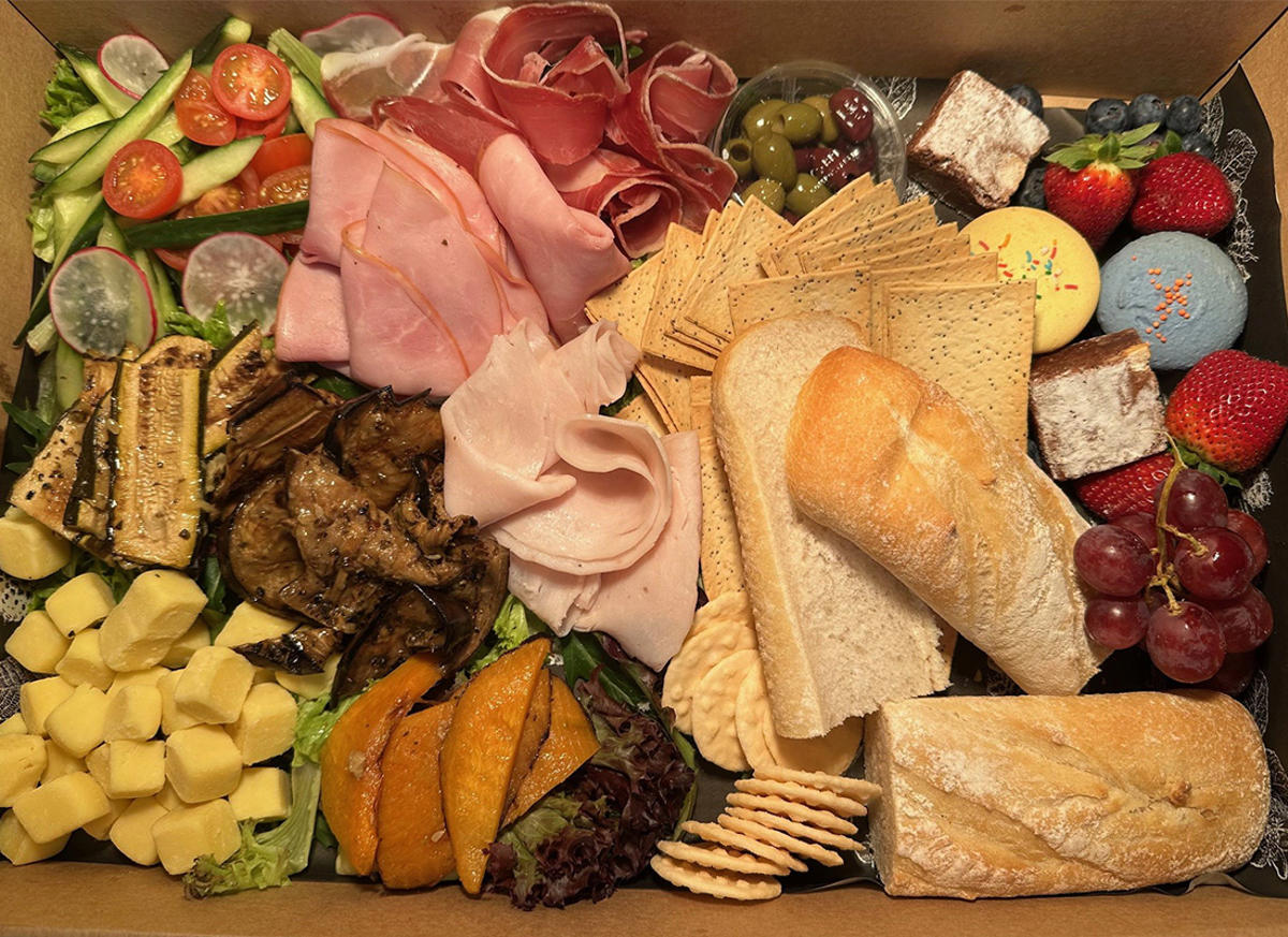 Hamper of assorted foods, including ham, turkey, cheese, zucchini, pastrami, tomato, olives, bread, cherries, strawberries, cookies and macarons.