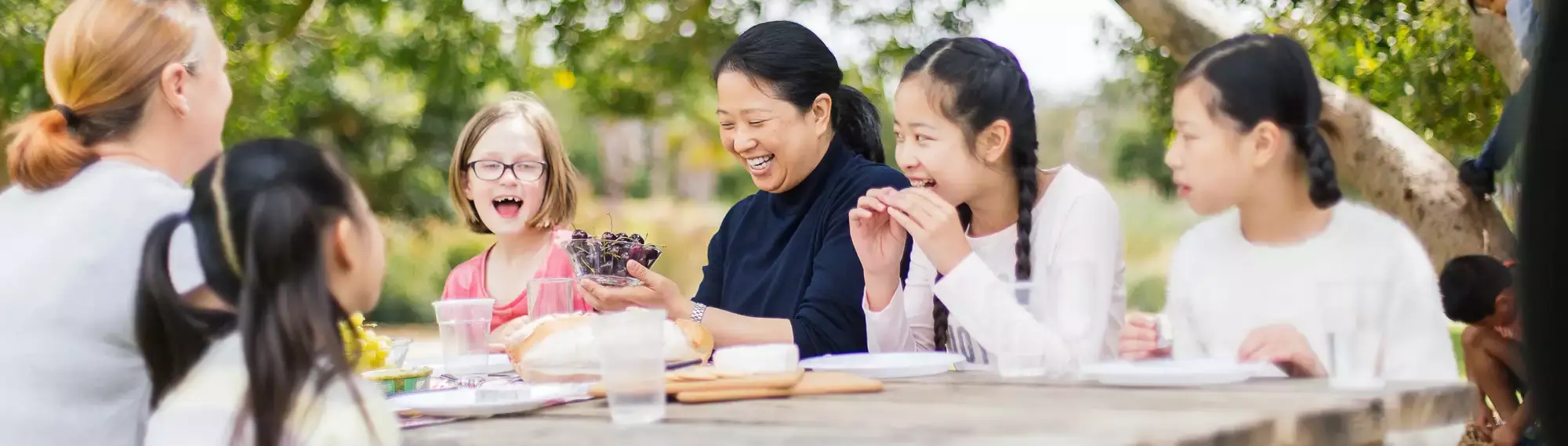 A group of adults and children are sitting at an outdoor table laughing. One woman is holding a bowl of cherries.