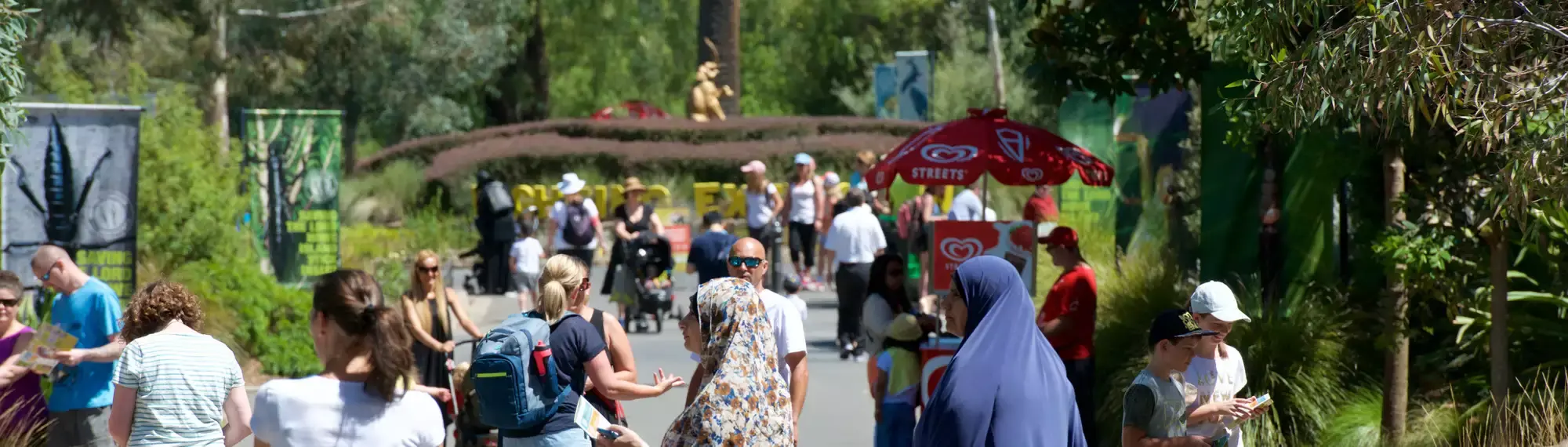 A view of the crowded Main Drive at Melbourne Zoo, with a red Streets ice cream cart in the mid-ground and the gold Elephant statue in the background.