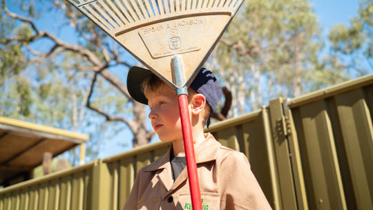 Junior "Zoo-Keeper" holding a rake, with red handle and dust-covered blue head, looking left and standing in front of a corrugated olive yellow fence.