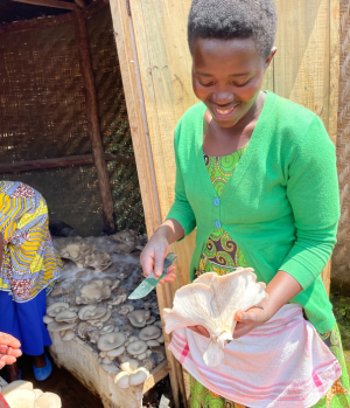 A women holds a large oyster mushroom and a knife in her hand; she is looking at the mushroom with a smile on her face,